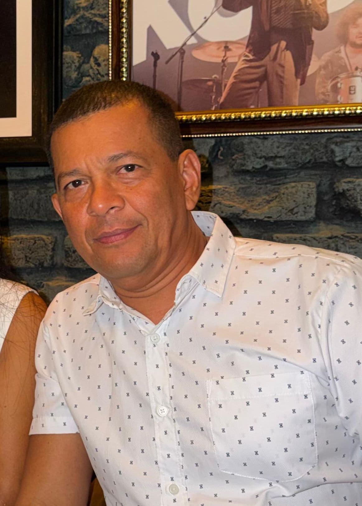 Hombre con camisa blanca de estampado pequeño sonriendo frente a una pared de ladrillos rústica.